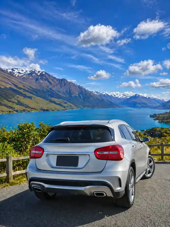 Silver SUV parked at a scenic lakeside viewpoint with snow-capped mountains and blue skies in the background, showcasing the beauty of South Island travel and car rentals services in Christchurch.